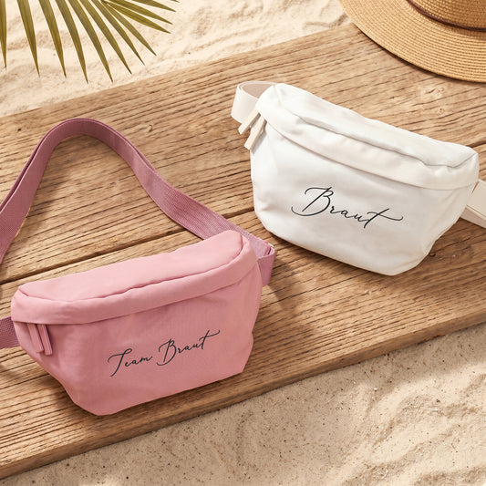 Rosa Bauchtasche mit Wunschtext Team Braut und cremefarbene Tasche mit Braut-Schriftzug auf Holzuntergrund