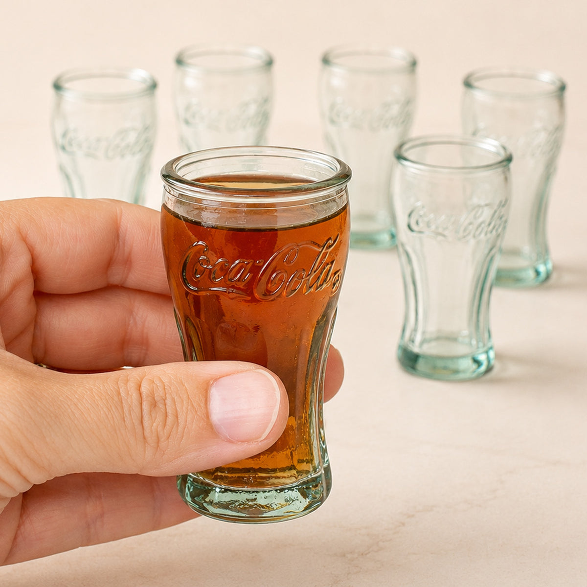 6 Schnapsgläser im Coca Cola Retro Design, eins ist gefüllt und wird von Hand gehalten