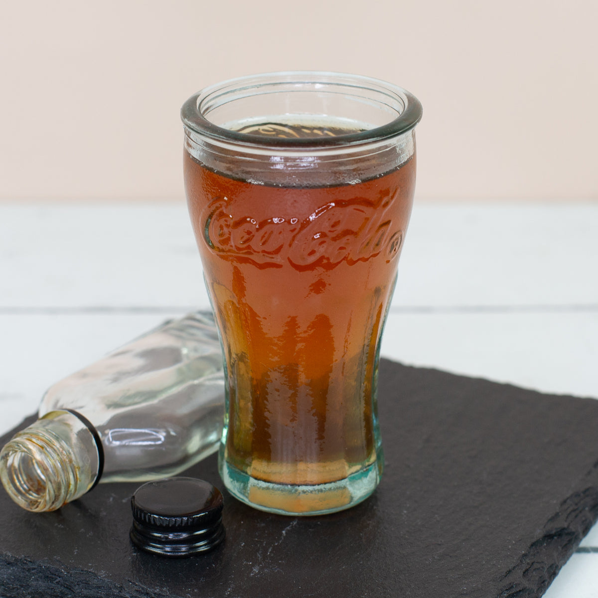 Rundliches Coca-Cola Stamperglas mit Getränk und kleiner Flasche auf dunkler Schieferplatte