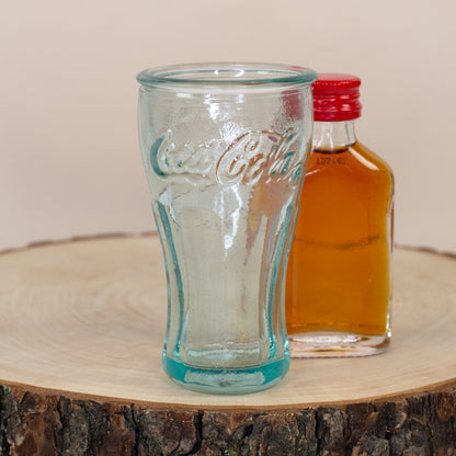 Leeres Coca-Cola Schnapsglas aus recyceltem Glas mit kleiner Flasche auf Holzscheibe