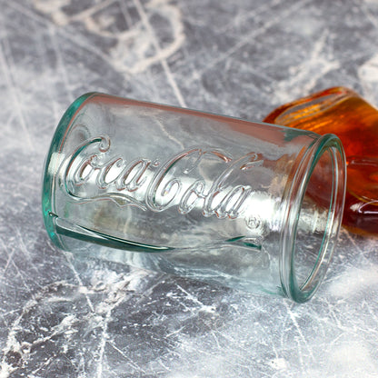 Liegendes Coca-Cola Shotglas aus recyceltem Glas mit geprägtem Schriftzug und Mini-Flasche im Hintergrund