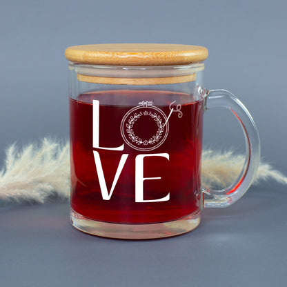 Glastasse mit rotem Tee, LOVE Stickmotiv und aufgesetztem Bambusdeckel vor grauem Hintergrund