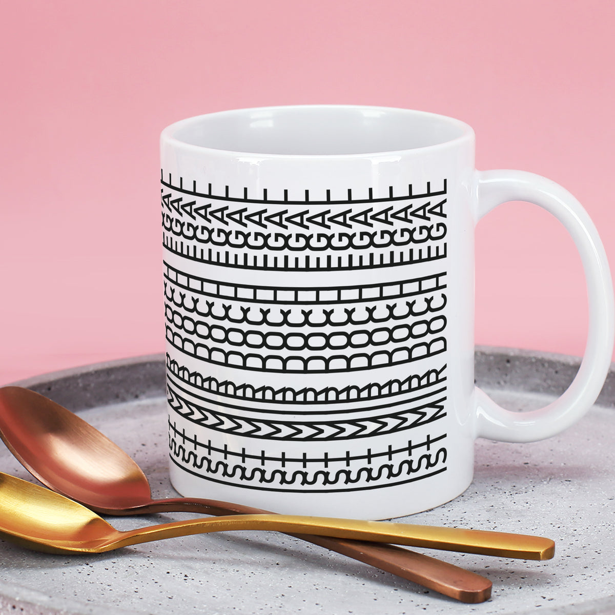 Weiße Tasse mit Schwarz-Weiß-Muster auf rosa Hintergrund neben goldenen Löffeln