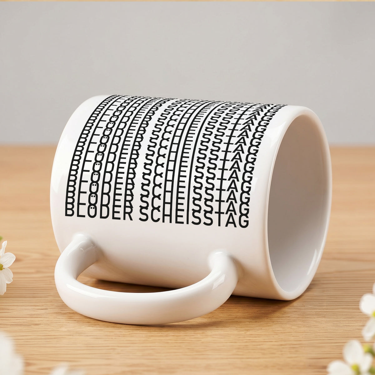 Weiße Kaffeetasse liegend auf hellem Holztisch mit versteckter Botschaft „Blöder Scheißtag“ im schwarzen Musterdruck