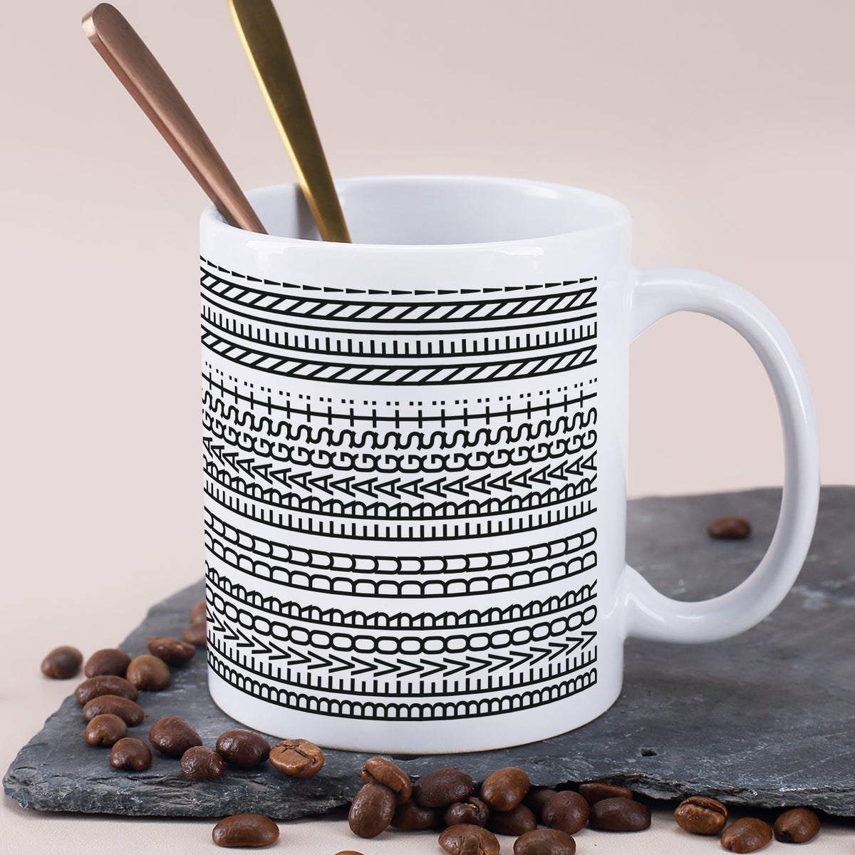 Weiße Keramiktasse mit schwarzem Musterdruck, Löffeln und Kaffeebohnen auf Schieferplatte
