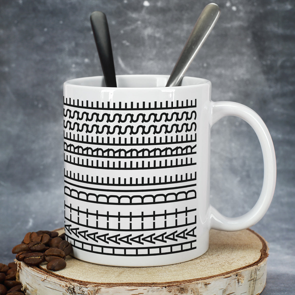 Keramiktasse mit grafischem Schwarz-Weiß-Muster, Löffeln und Kaffeebohnen auf Holzscheibe
