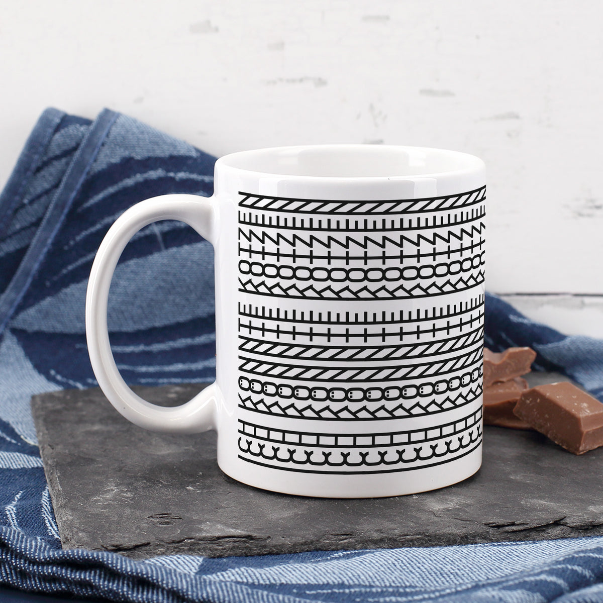 Kaffeetasse mit grafischem Schwarz-Weiß-Muster auf Schieferplatte neben blauem Tuch und Schokolade