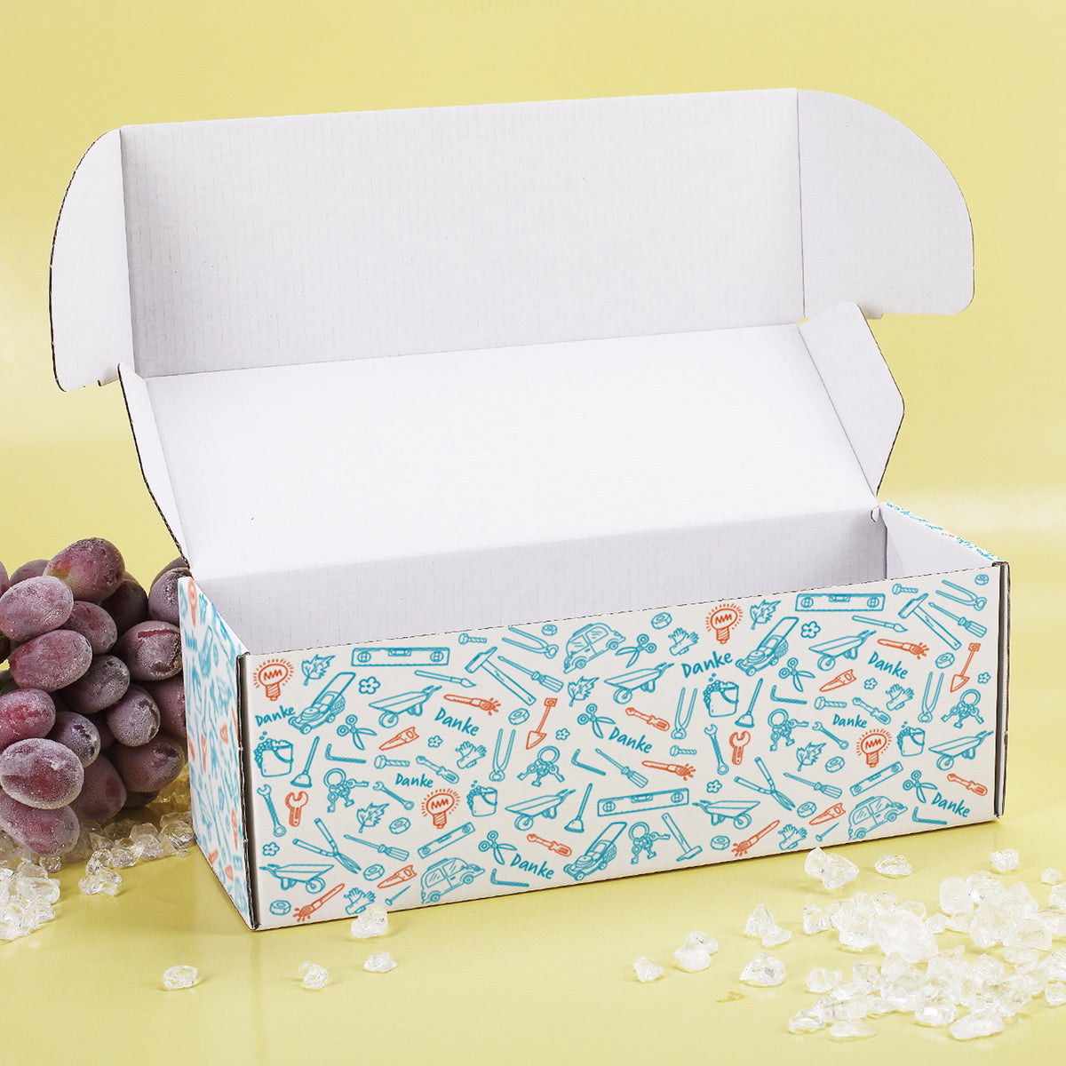 Geöffnete Geschenkbox mit farbigem Werkzeug-Muster und Deko auf gelbem Hintergrund mit WeintraubenDeko.