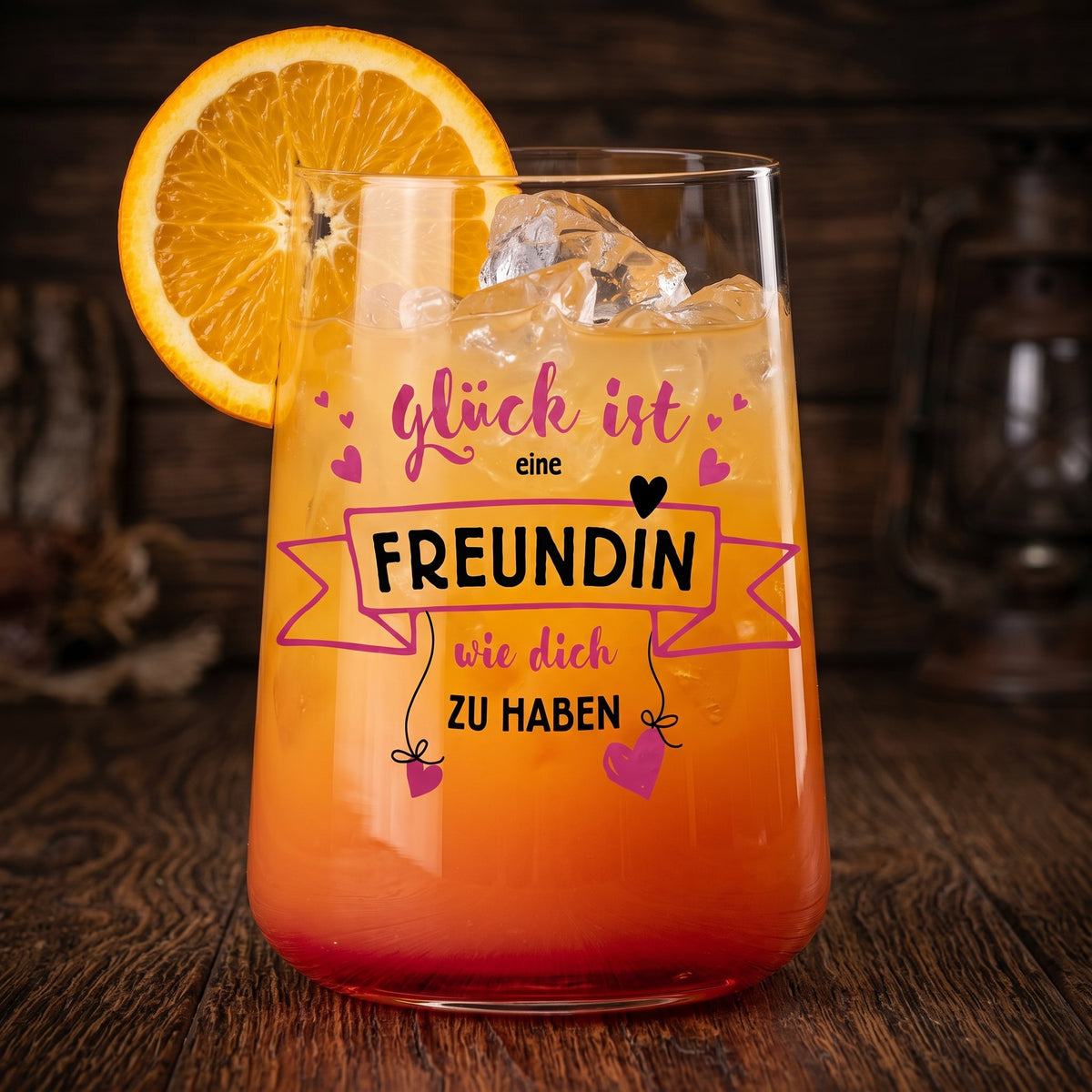 Trinkglas mit orangefarbenem Cocktail, Eiswürfeln, Orangenscheibe und Spruch „Glück ist eine Freundin wie dich zu haben“