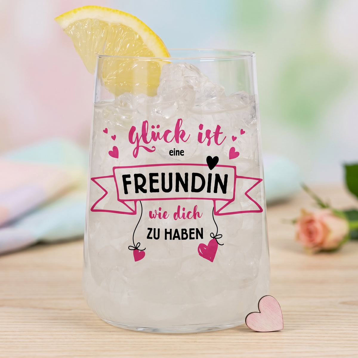 Trinkglas mit Eiswürfeln, Zitronenscheibe und Freundinnen-Spruch auf hellem Tisch mit pastelligem Hintergrund