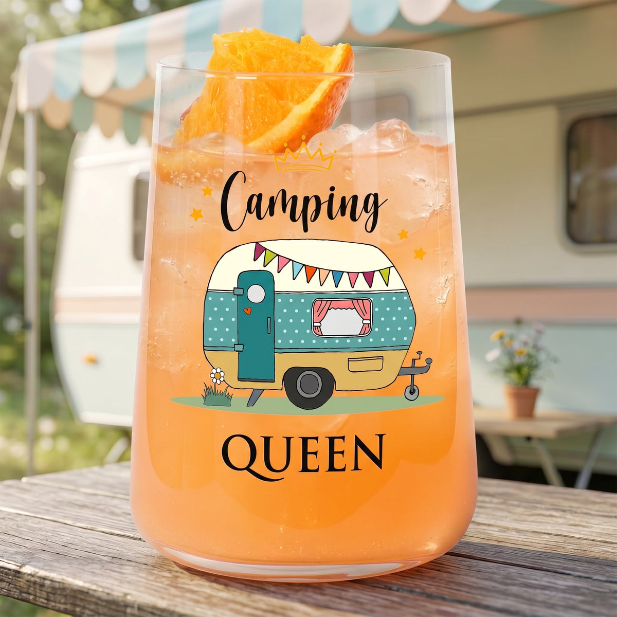 Trinkglas mit Camping Queen Wohnwagen-Motiv vor Wohnwagen-Hintergrund und orangefarbenem Getränk