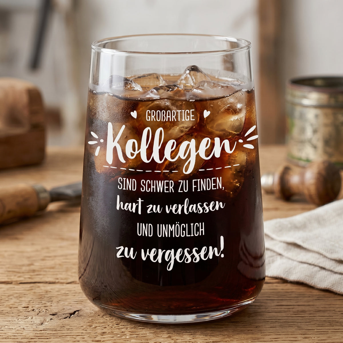 Trinkglas mit Kollegen Spruch, gefüllt mit dunklem Kaltgetränk und Eiswürfeln auf Holztisch