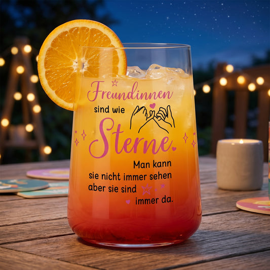 Trinkglas für Freundinnen mit rosa-schwarzem Spruch „Freundinnen sind wie Sterne“ gefüllt mit orangem Getränk und Orangenscheibe auf einem Tisch bei Abendstimmung