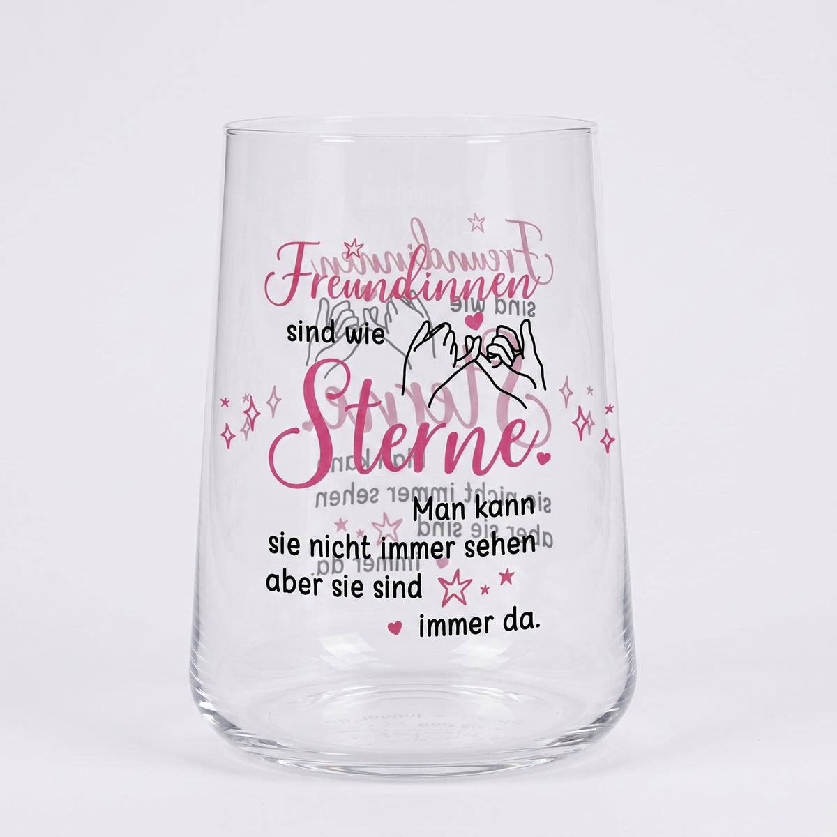 Leeres Trinkglas mit rosa-schwarzem Motivdruck „Freundinnen sind wie Sterne“ und Illustration zweier Hände auf weißem Hintergrund