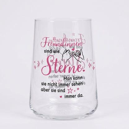 Leeres Trinkglas mit rosa-schwarzem Motivdruck „Freundinnen sind wie Sterne“ und Illustration zweier Hände auf weißem Hintergrund