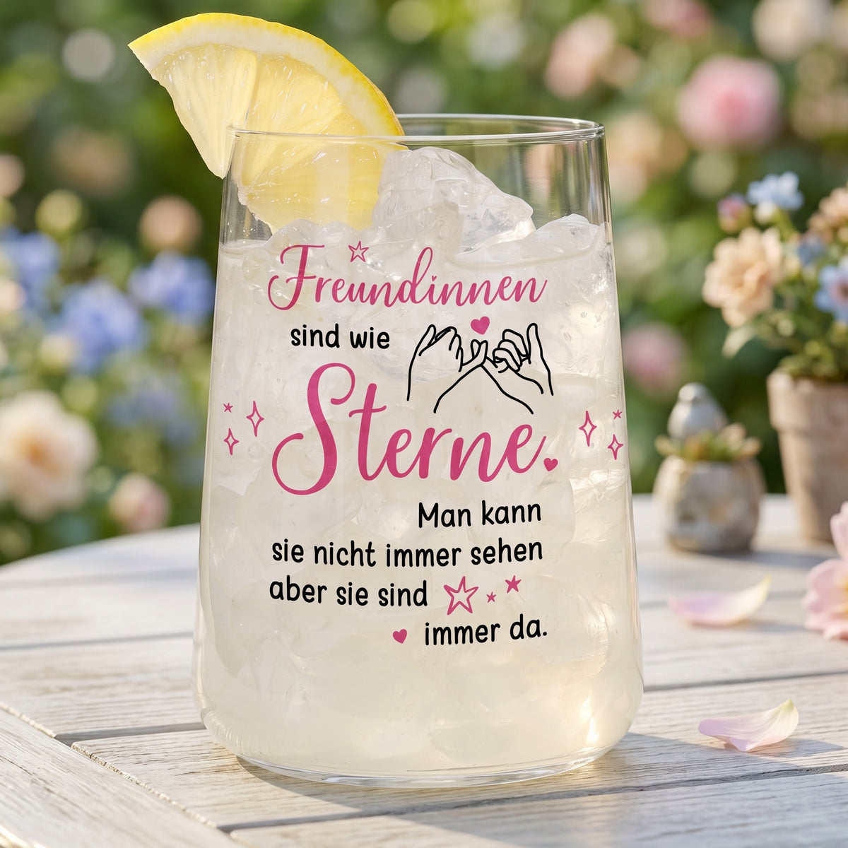Trinkglas mit Spruch für Freundinnen gefüllt mit Eis und Zitronenscheibe auf einem Tisch vor blumigem Hintergrund