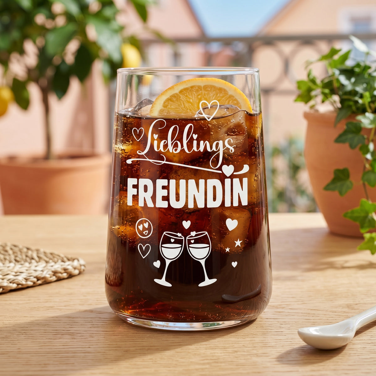 Trinkglas mit Aufdruck Lieblingsfreundin, gefüllt mit dunklem Getränk und Eiswürfeln, garniert mit Orangenscheibe, auf Holztisch im hellen Ambiente