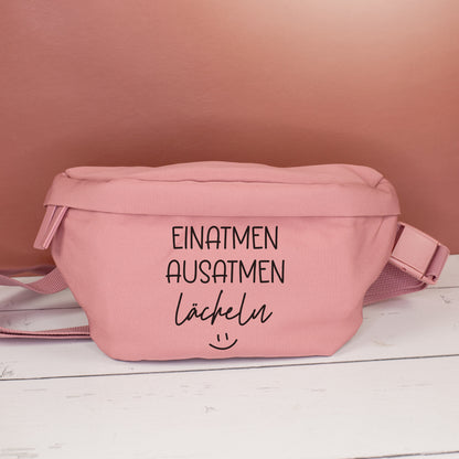 Rosafarbene Bauchtasche mit Spruch Einatmen Ausatmen Lächeln frontal auf hellem Holztisch vor rötlicher Wand