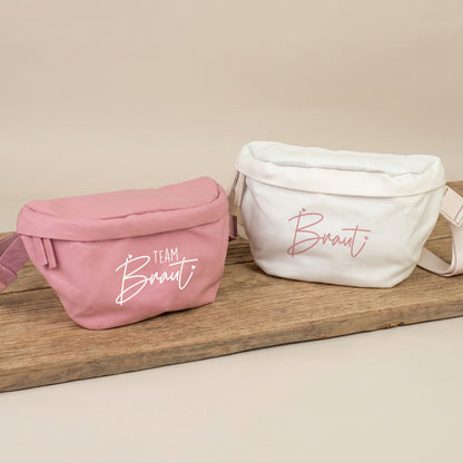 Rosa JGA Bauchtasche mit Aufdruck Team Braut und cremefarbene Bauchtasche mit Aufdruck Braut auf Holzbrett