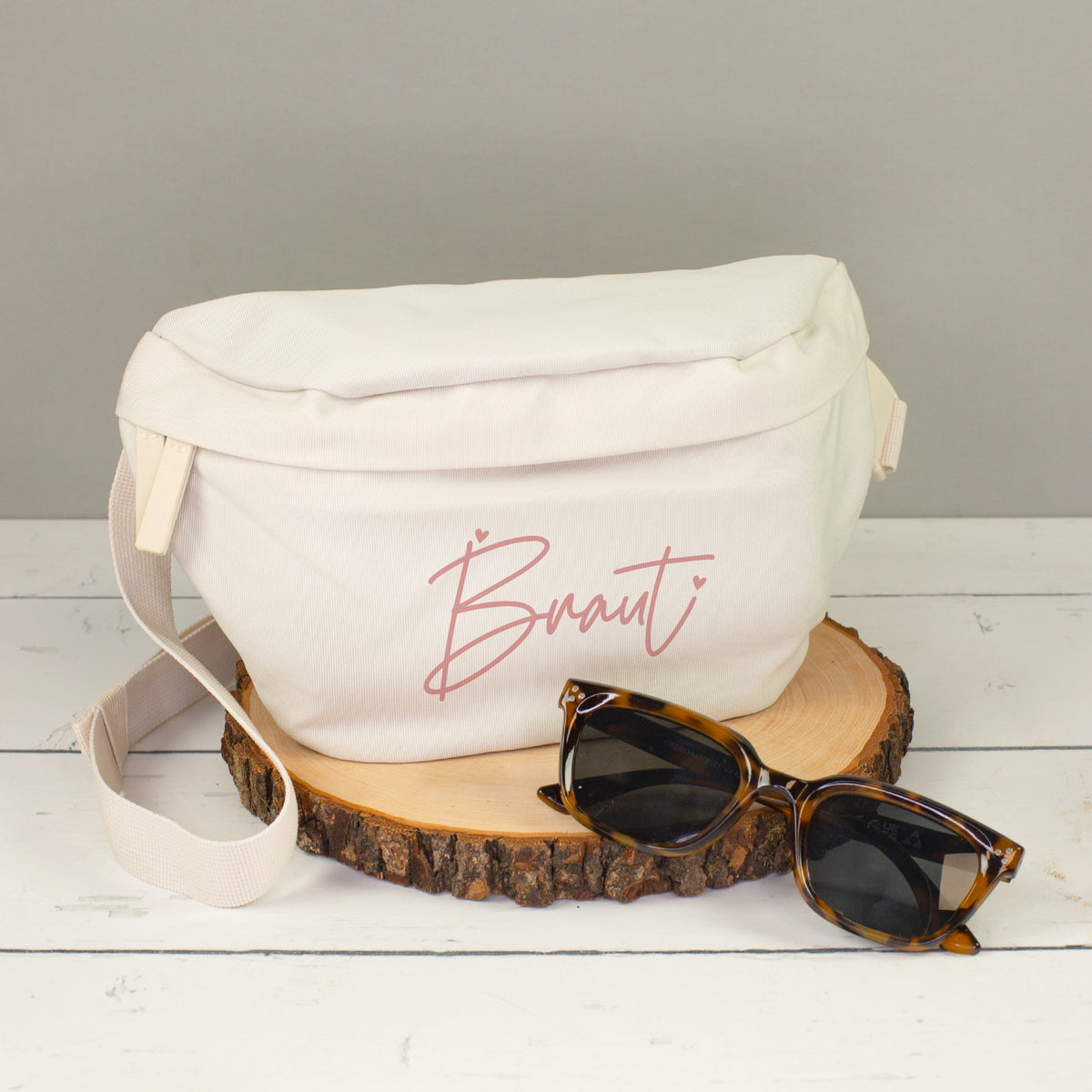 Cremefarbene Bauchtasche mit rosa Braut-Aufdruck auf Holzscheibe, dekoriert mit Sonnenbrille