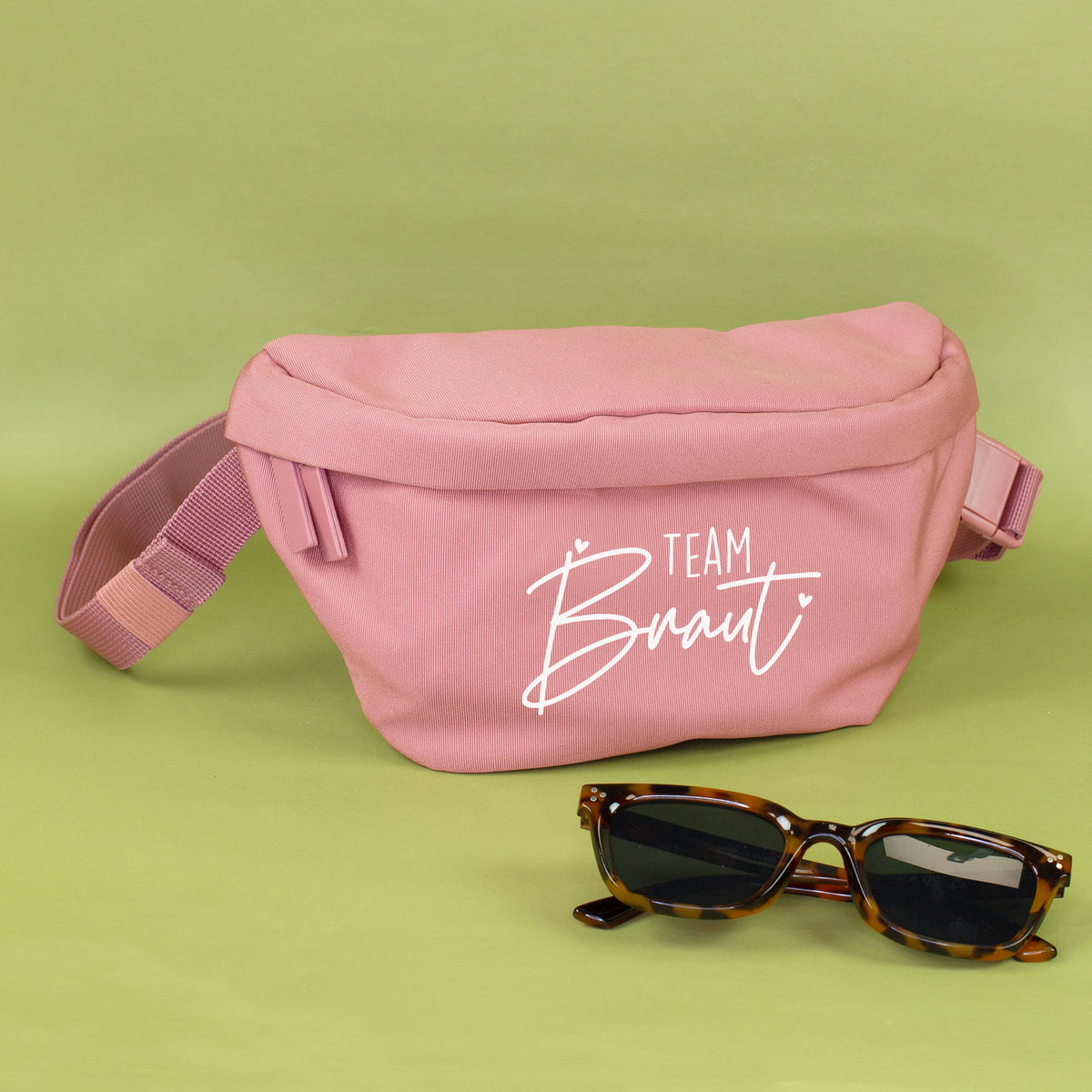 Rosa Bauchtasche mit weißem Team Braut Aufdruck auf grünem Hintergrund, dekoriert mit Sonnenbrille
