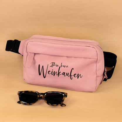 Rosa Bauchtasche mit Spruch Bin kurz Weinkaufen und schwarzem Gurt auf weißem Hintergrund