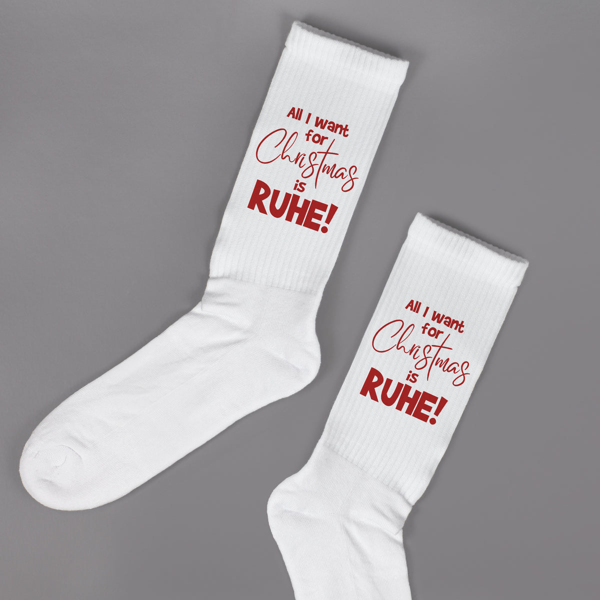 Einzelne weiße Sportsocken mit rotem Weihnachtsspruch ‚All I want for Christmas is RUHE!‘ vor grauem Hintergrund.