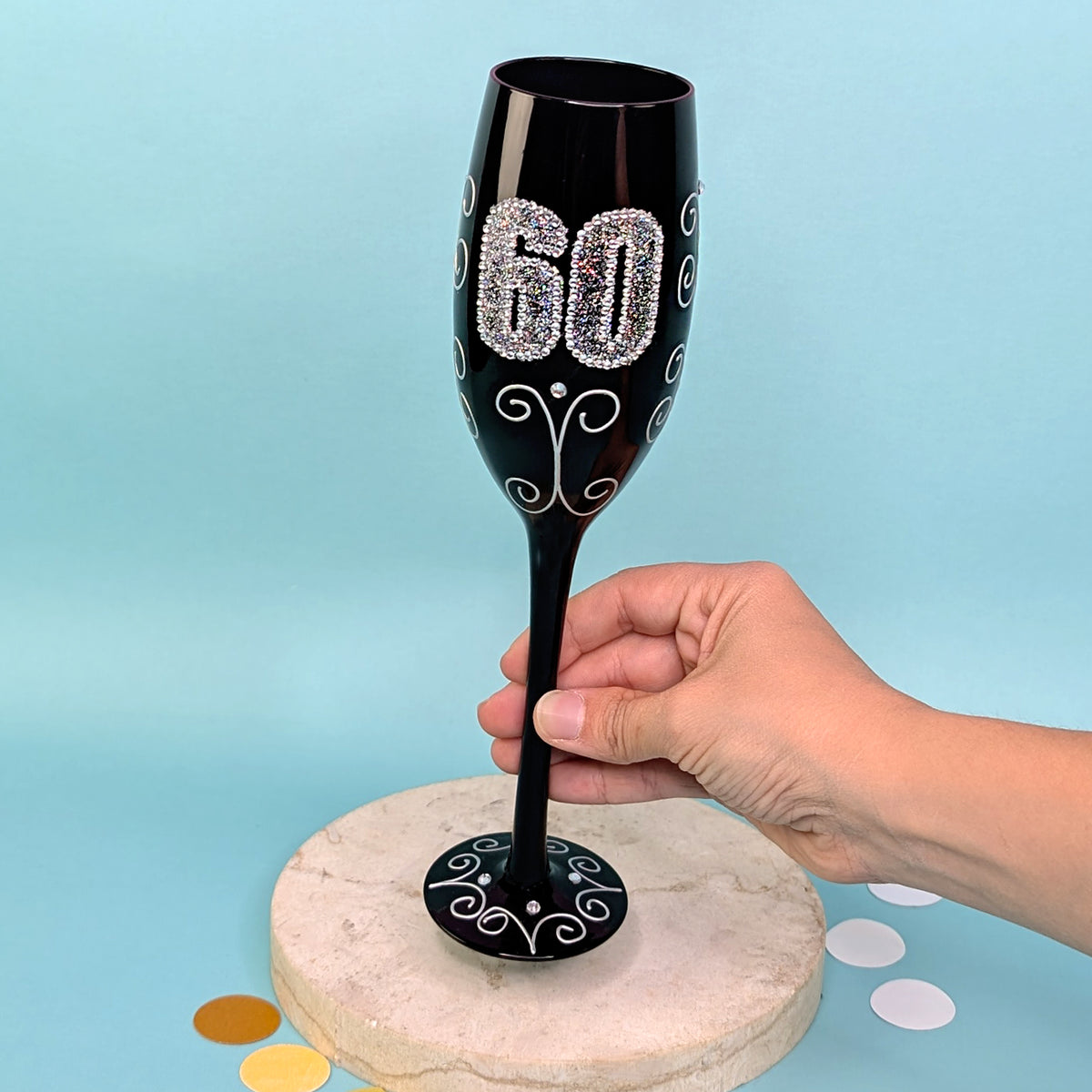 Schwarzes Sektglas mit glitzernder 60 und Ornamenten, frontal fotografiert, gehalten von einer Hand