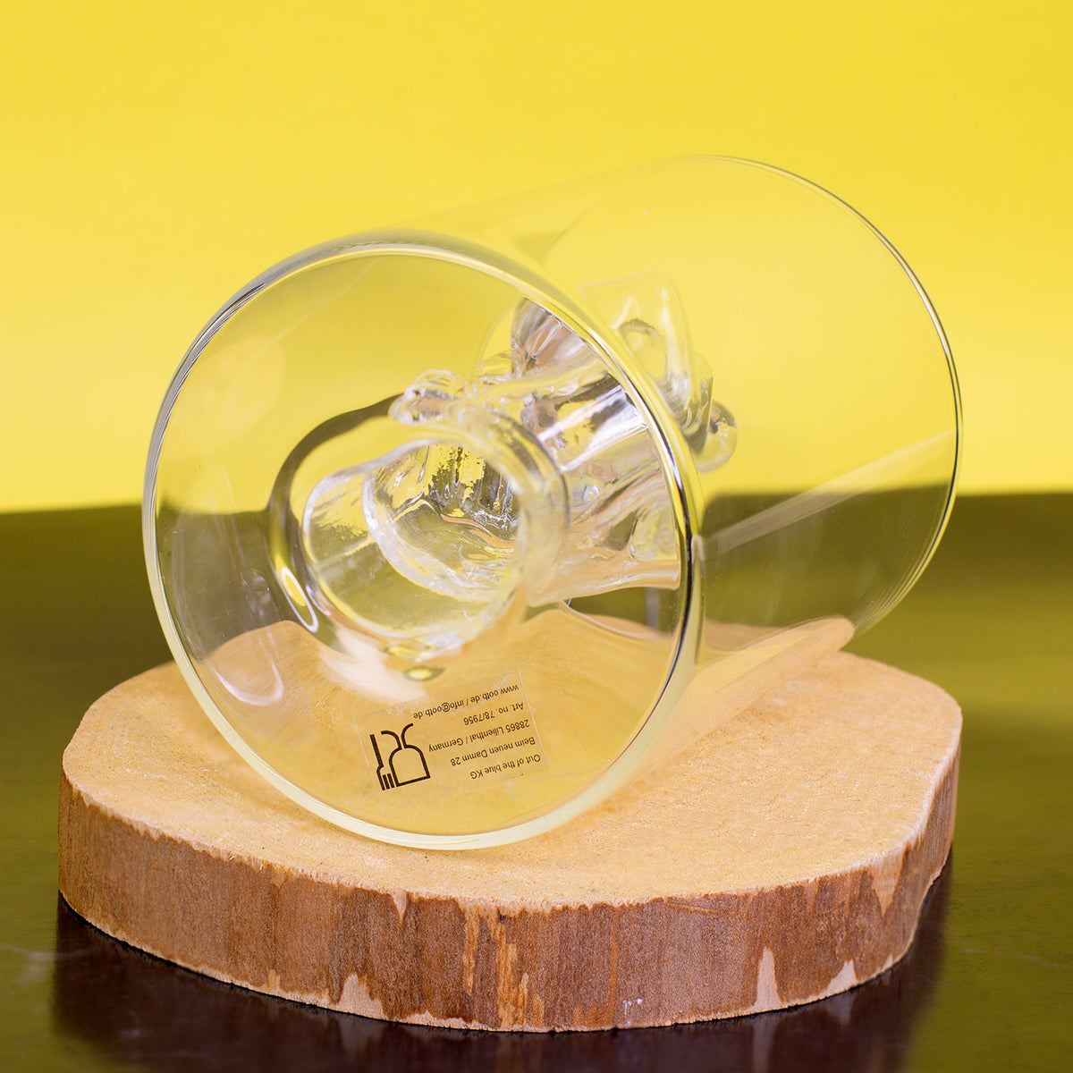 Trinkglas mit Mittelfinger Figur liegend auf Holzscheibe vor gelbem Hintergrund