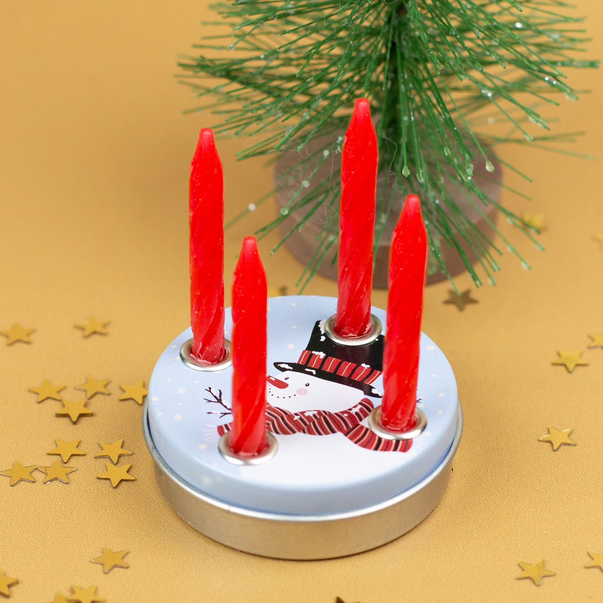 Mini-Adventskranz mit vier eingesetzten roten Kerzen im Schneemann-Design, vor einem kleinen Weihnachtsbaum und goldenen Stern-Konfetti.