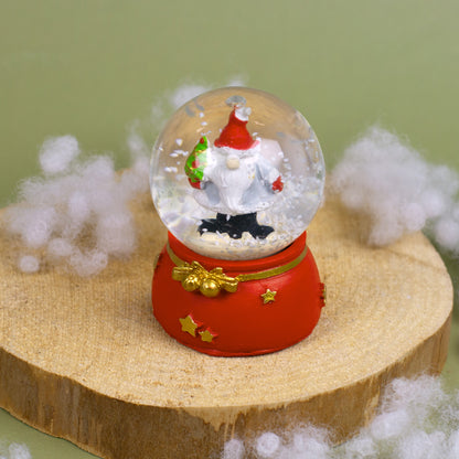 Schneekugel mit Weihnachtswichtel und geschmücktem Tannenbaum, auf Holzpodest mit Kunstschnee