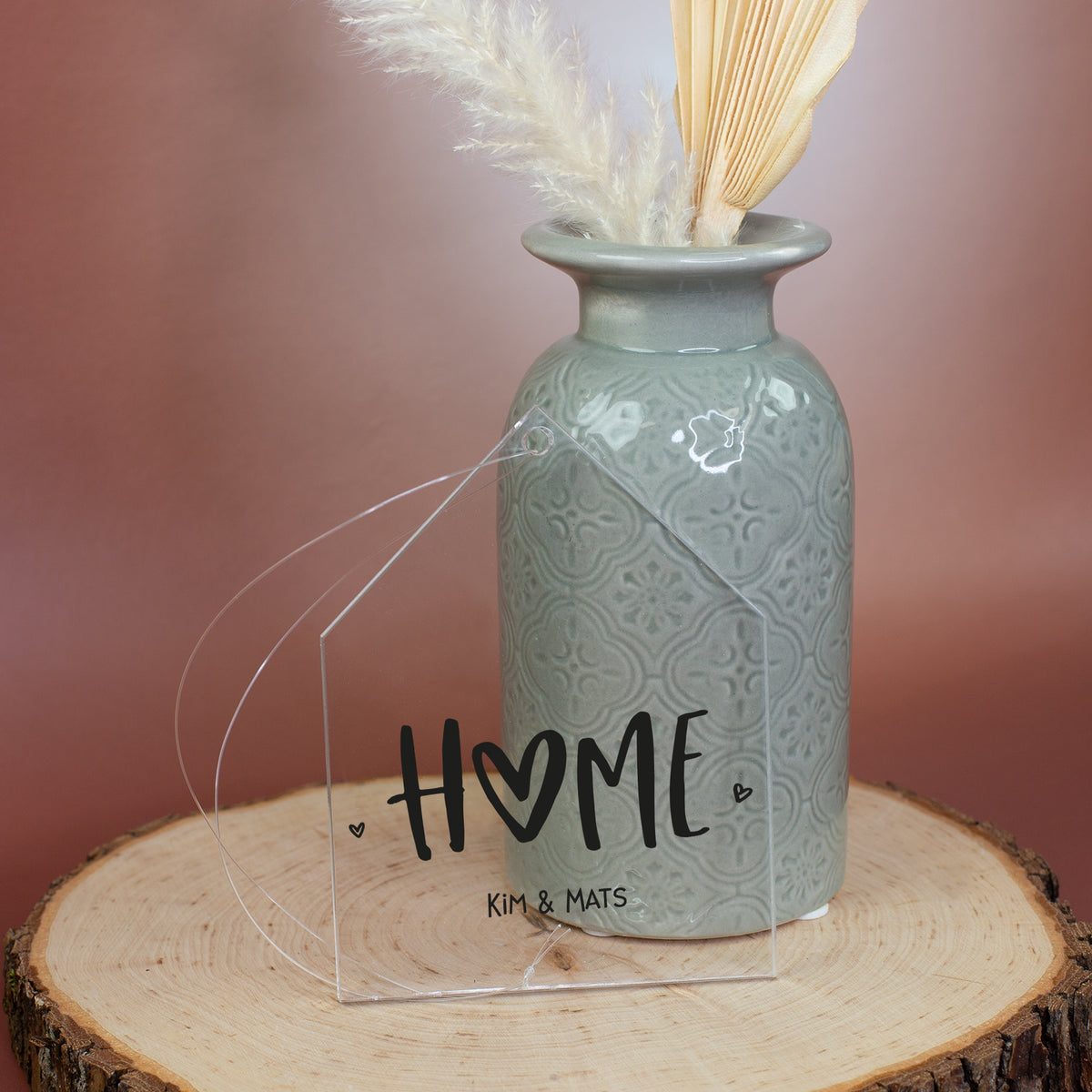 Deko-Anhänger aus Acryl in Hausform mit HOME-Aufdruck und Wunschtext Kim und Mats neben grauer Vase auf Holzscheibe