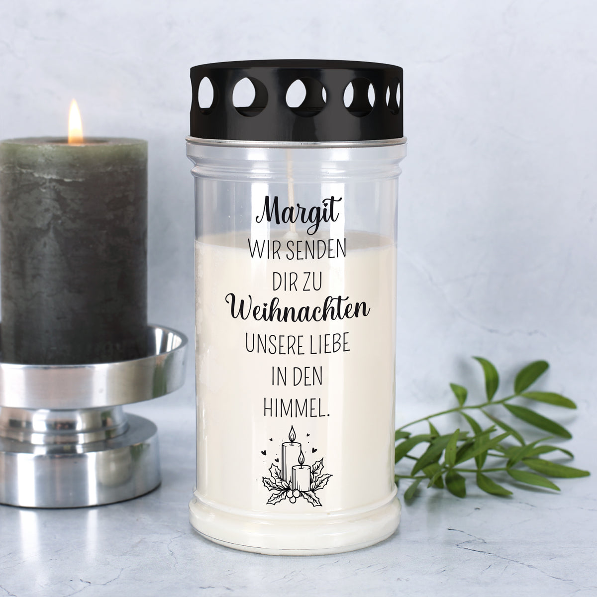 Erinnerungs-Grablicht mit Name „Margit“ und Weihnachtsmotiv, daneben brennende Kerze und grüner Zweig