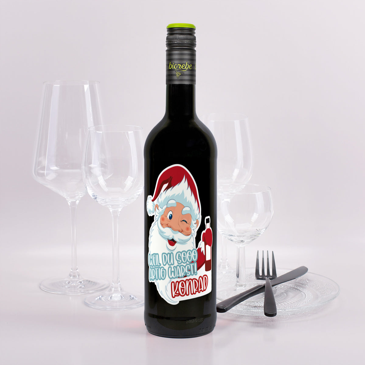 Weinflasche mit rotem Wein, Etikett Weihnachtsmann-Motiv „Weil du sooo artig warst! Konrad“, festlich dekoriert mit Gläsern.