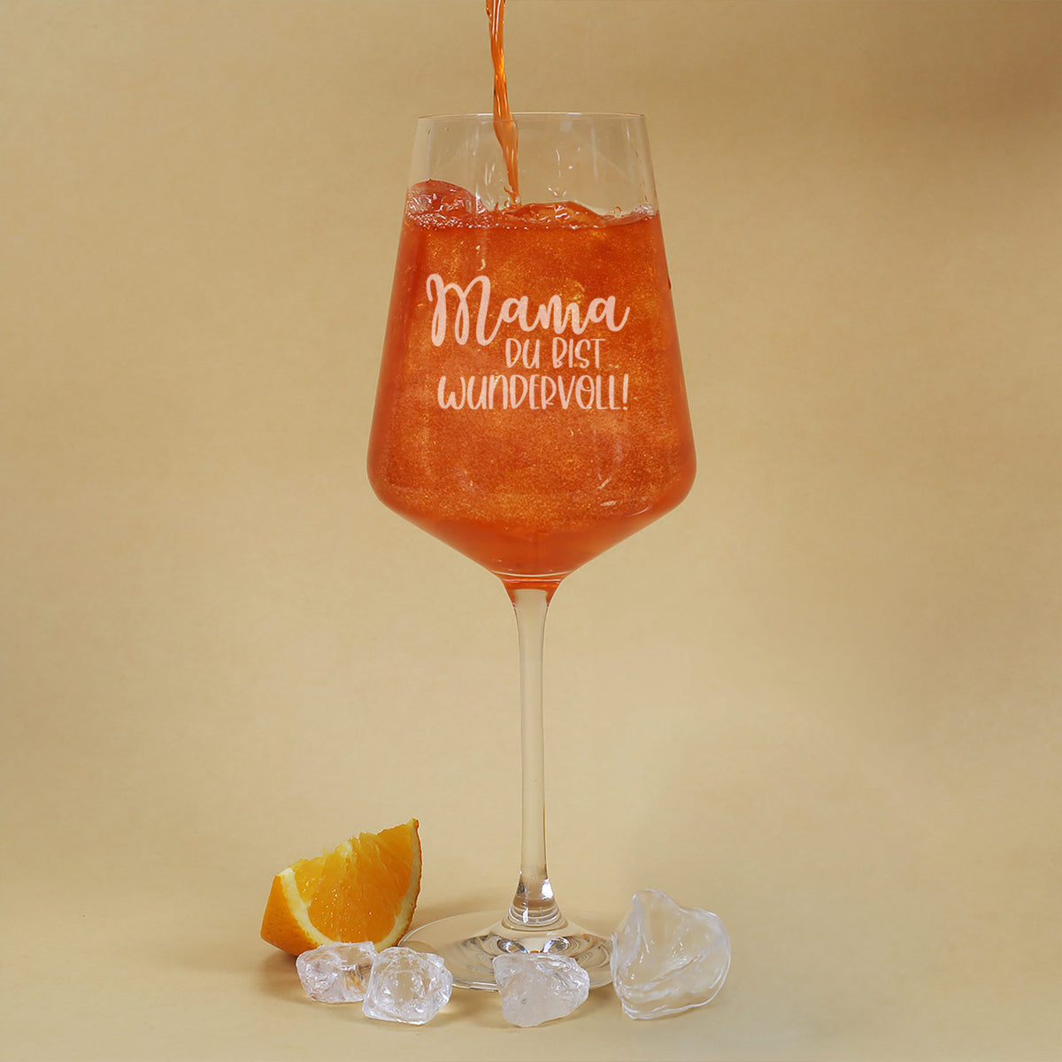 Graviertes Cocktailglas mit Glittergetränk wird befüllt, daneben Eiswürfel und Orangenschnitz auf gelbem Hintergrund