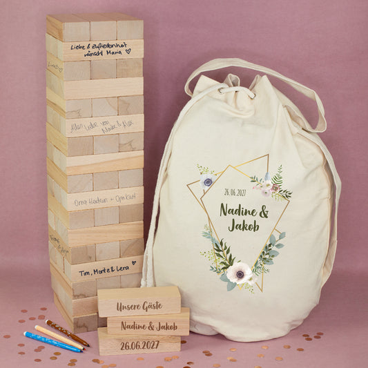 XXL Wackelturm aus Holz als Hochzeitsgästebuch mit beschrifteten Steinen, personalisiertem Seesack mit Blumenmotiv sowie drei gravierten Holzsteinen vor rosafarbenem Hintergrund