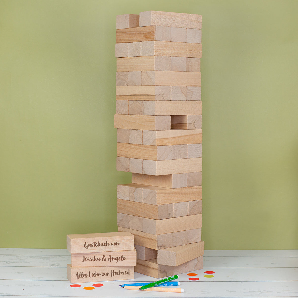 Großer Holz Wackelturm als Hochzeitsgästebuch mit drei gravierten Beispielsteinen für Namen und Hochzeitsdatum vor grünem Hintergrund