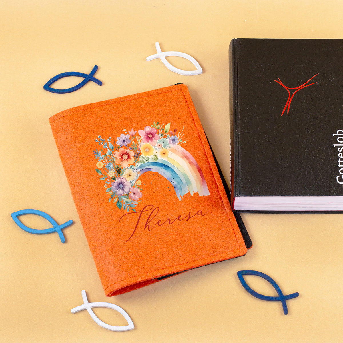 Personalisierte Gotteslobhülle aus orangefarbenem Filz mit Regenbogen, Blumen und Name neben Gesangbuch