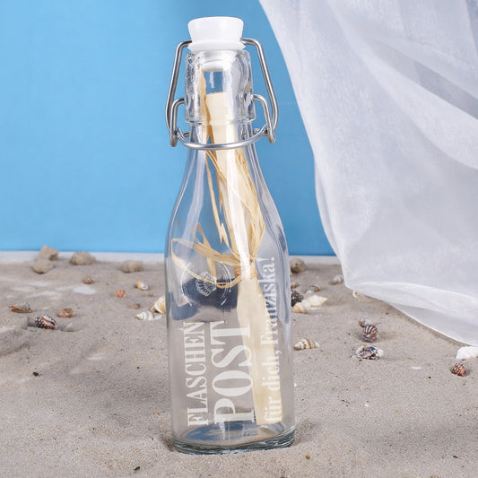 Dekorative Flaschenpost mit personalisiertem Text am Strand mit Muscheln und Stoff im Hintergrund