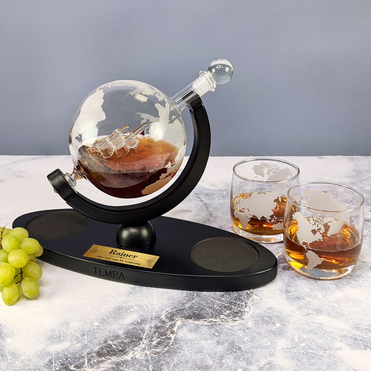 Whiskyset mit Weltkugel-Dekanter, Gläsern mit Weltkarten-Gravur und Trauben auf weiß-marmorierter Fläche