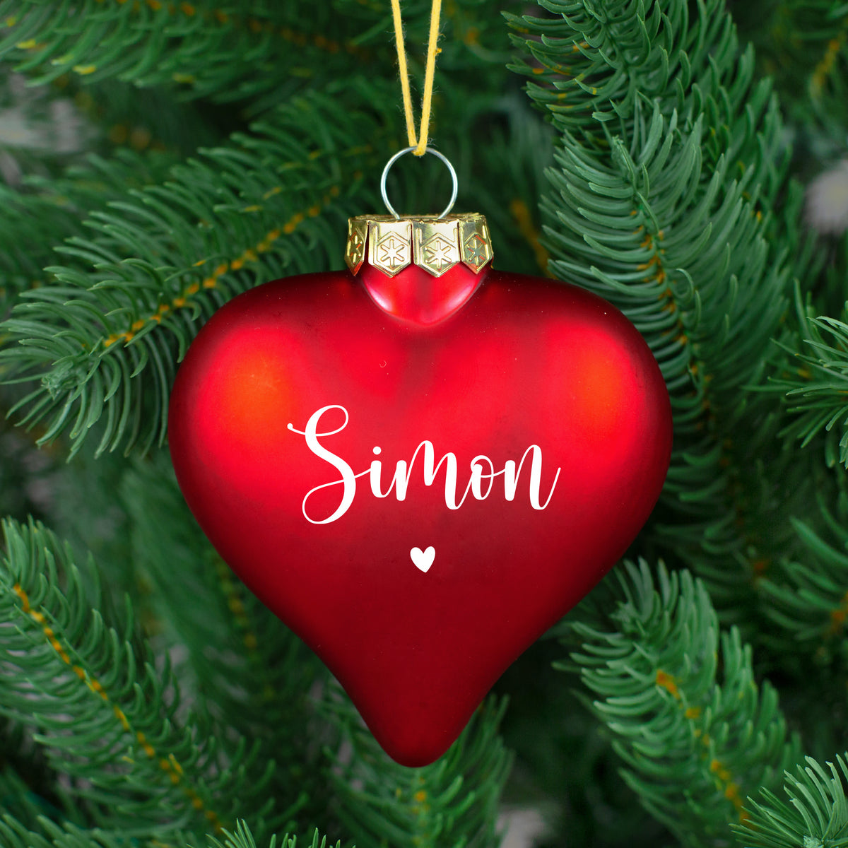 Rote, matte Herz-Christbaumkugel mit weißem Namen „Simon“ und kleinem Herz, an grünem Weihnachtsbaum.