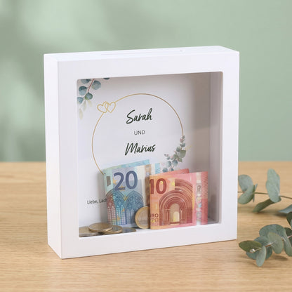 Bilderrahmen-Spardose für Sarah und Marius mit Geldscheinen und Münzen als kreatives Hochzeitsgeschenk auf Holztisch