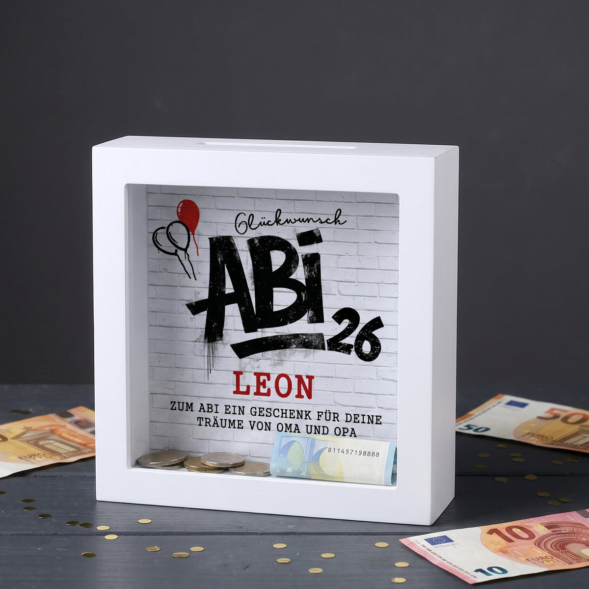 Weiße Bilderrahmen Spardose mit Abi 26 Motiv, Name Leon und Geldscheinen auf dunklem Tisch
