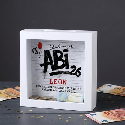 Weiße Bilderrahmen Spardose mit Abi 26 Motiv, Name Leon und Geldscheinen auf dunklem Tisch