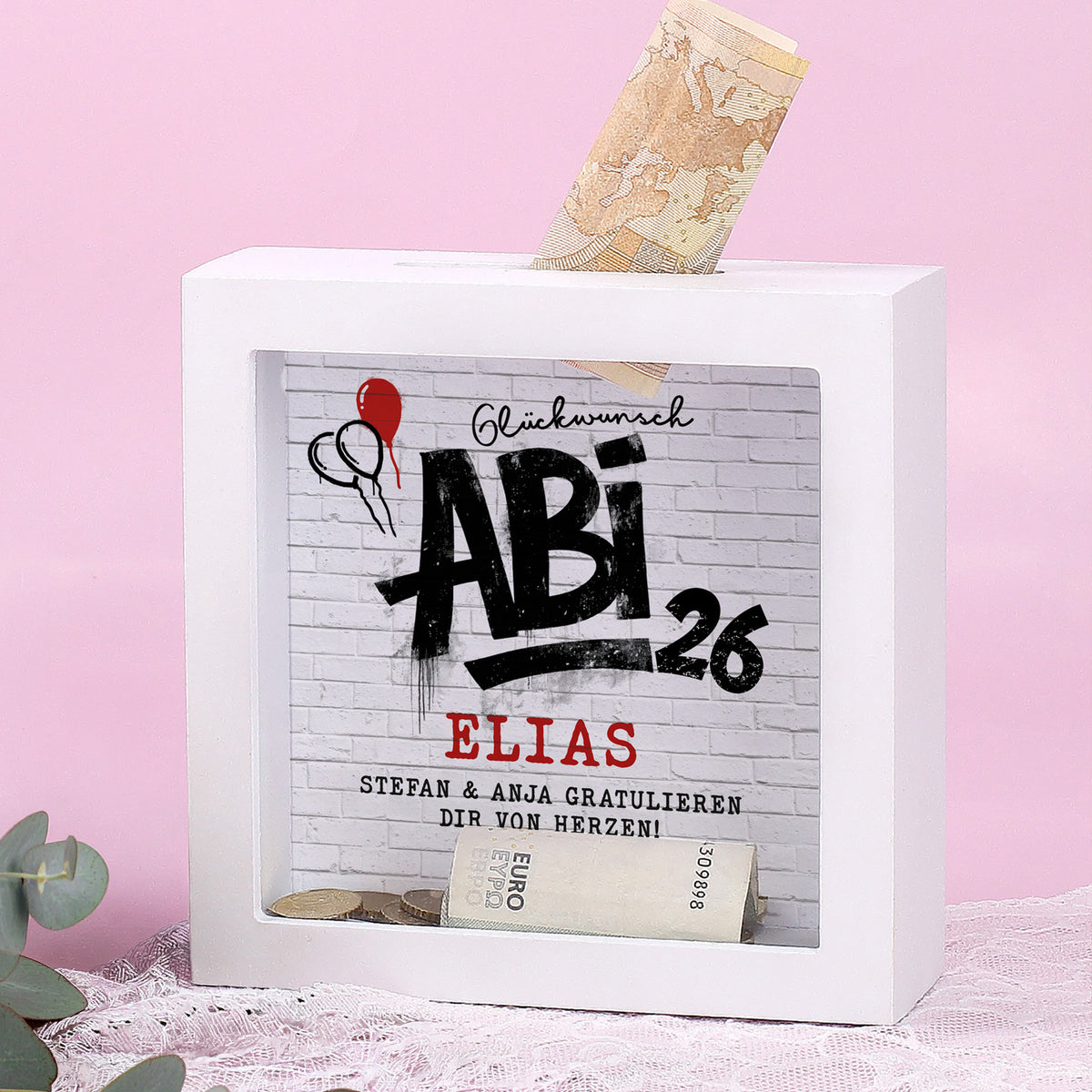 Spardose zum Abi 2026 mit Name Elias und Wunschtext vor rosa Hintergrund mit eingestecktem Geldschein