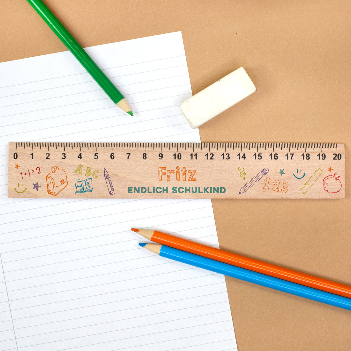 Holzlineal mit Name Fritz und Text „Endlich Schulkind“ auf Schulheft mit Buntstiften und Radiergummi