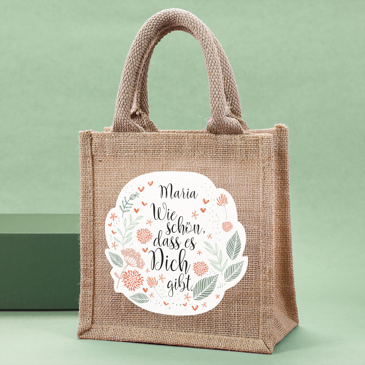 Geschenktasche aus Jute mit floralem Design, dem Spruch „Wie schön, dass es dich gibt.“ und personalisiertem Namen, seitlich fotografiert.