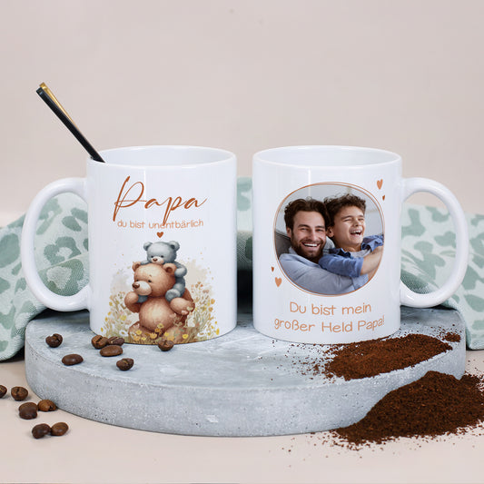 Zwei weiße Fototassen auf grauem Podest, links die Tasse mit Bärenmotiv „Papa du bist unentbärlich“, rechts die Rückseite mit Foto und dem Text „Du bist mein großer Held Papa!“.