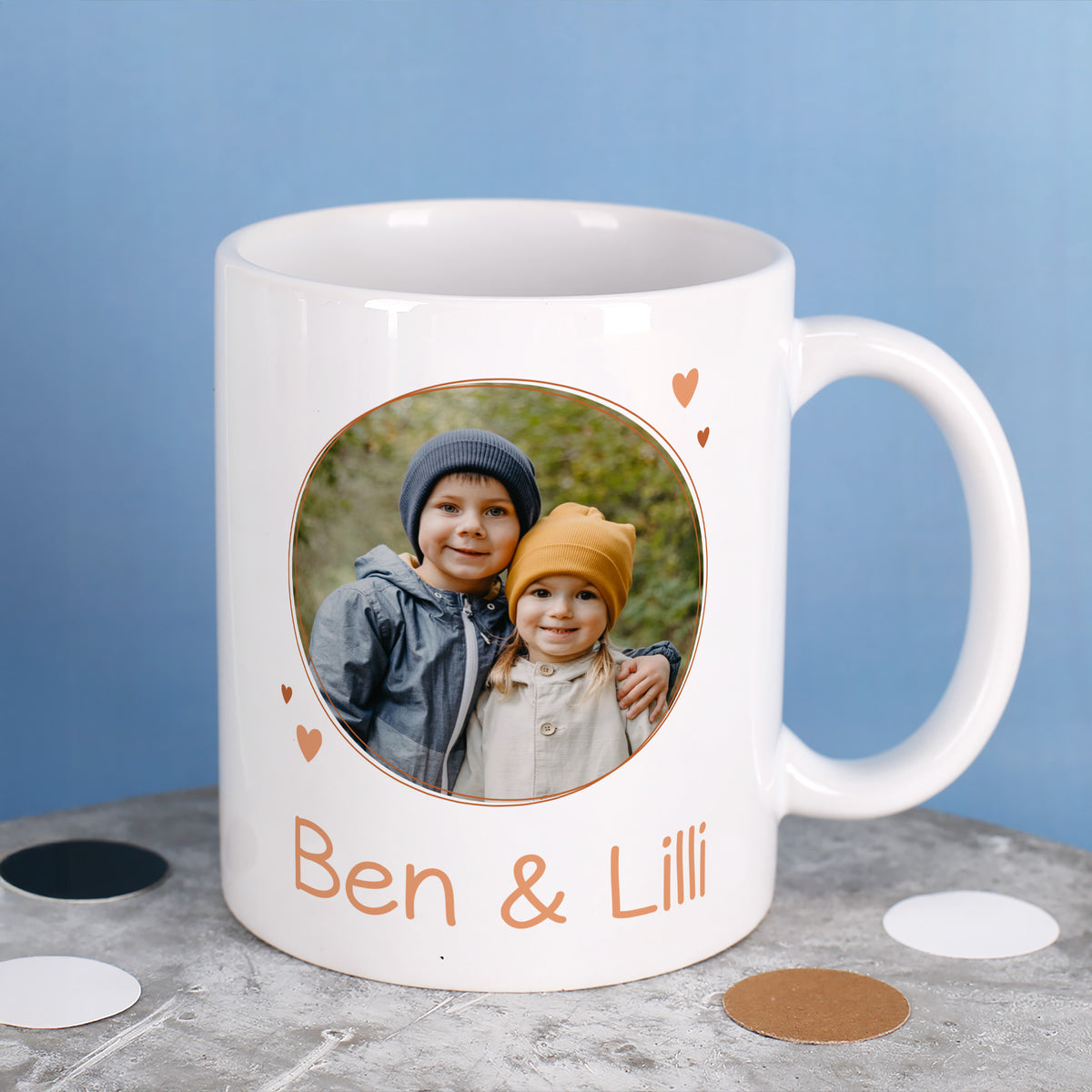 Weiße Fototasse mit rundem Kinderfoto und personalisiertem Text „Ben & Lilli“ auf grauem Untergrund.
