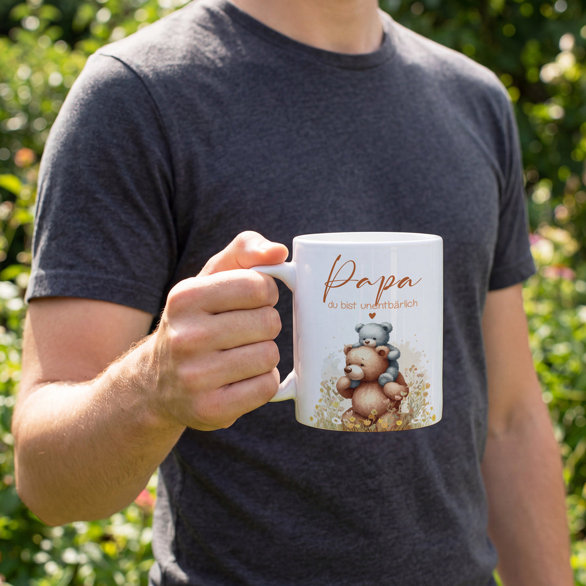Ein Mann hält die weiße Tasse mit Bärenmotiv „Papa du bist unentbärlich“ in der Hand im Garten.